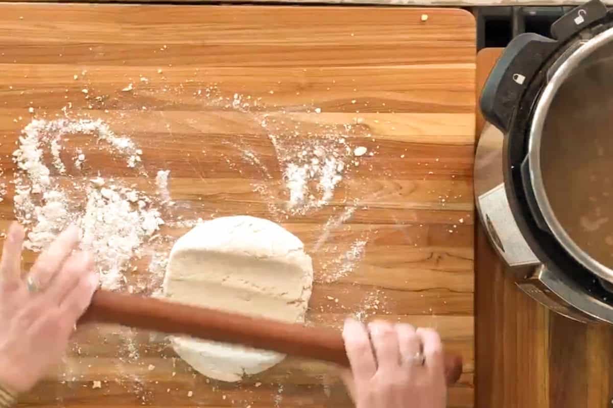 Hands rolling out dough with a wooden rolling pin on a floured wooden surface, next to a pressure cooker on the right side of the image.