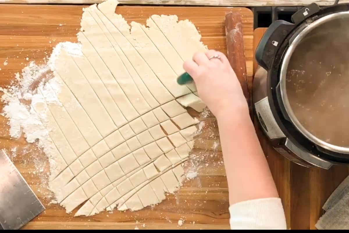 A person uses a green pastry cutter to slice rolled-out dough into strips on a floured wooden surface, next to a pressure cooker and a metal bench scraper.