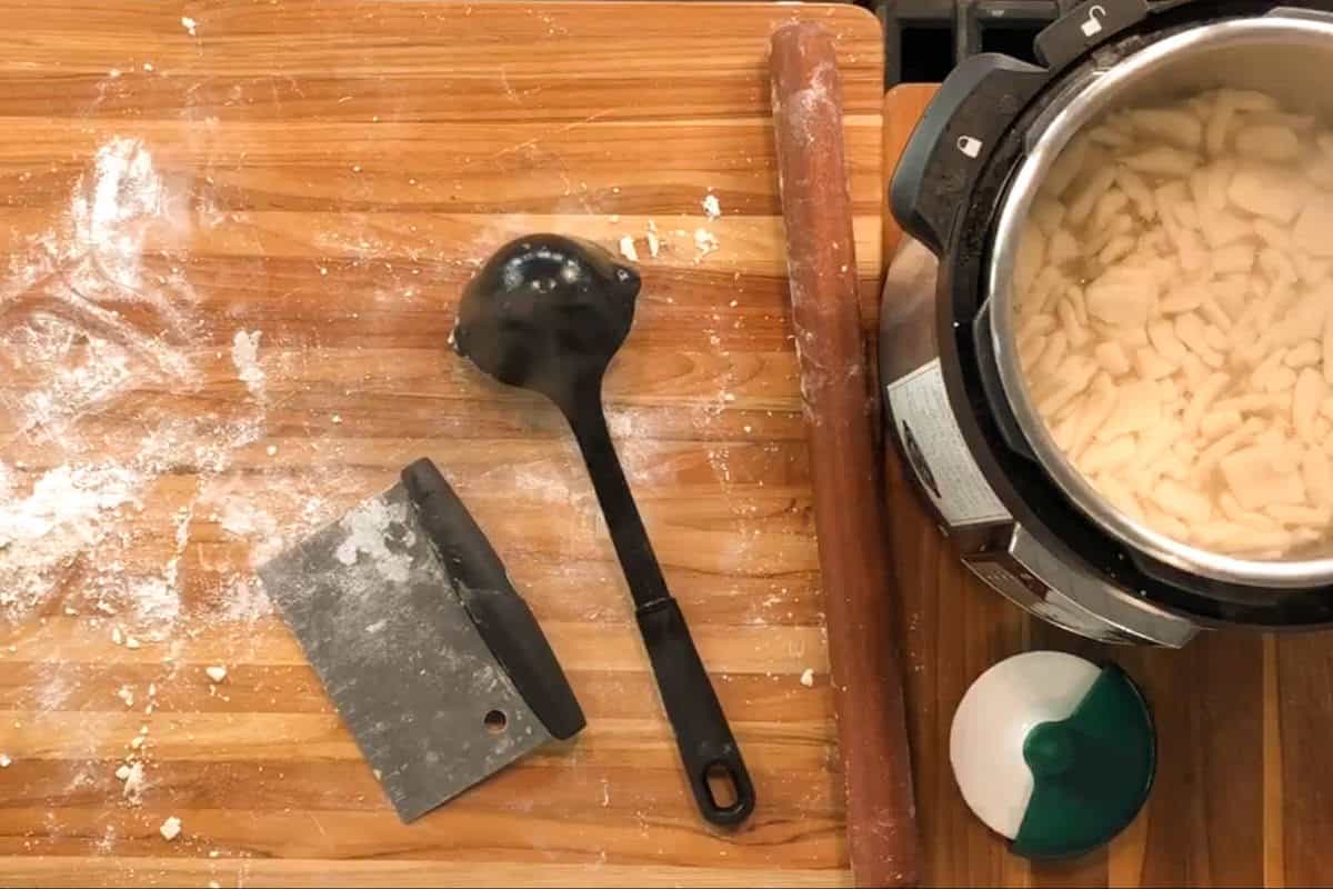 A wooden countertop with scattered flour, a metal bench scraper, a black soup ladle, a rolling pin, and an electric pressure cooker containing diced potatoes in water.