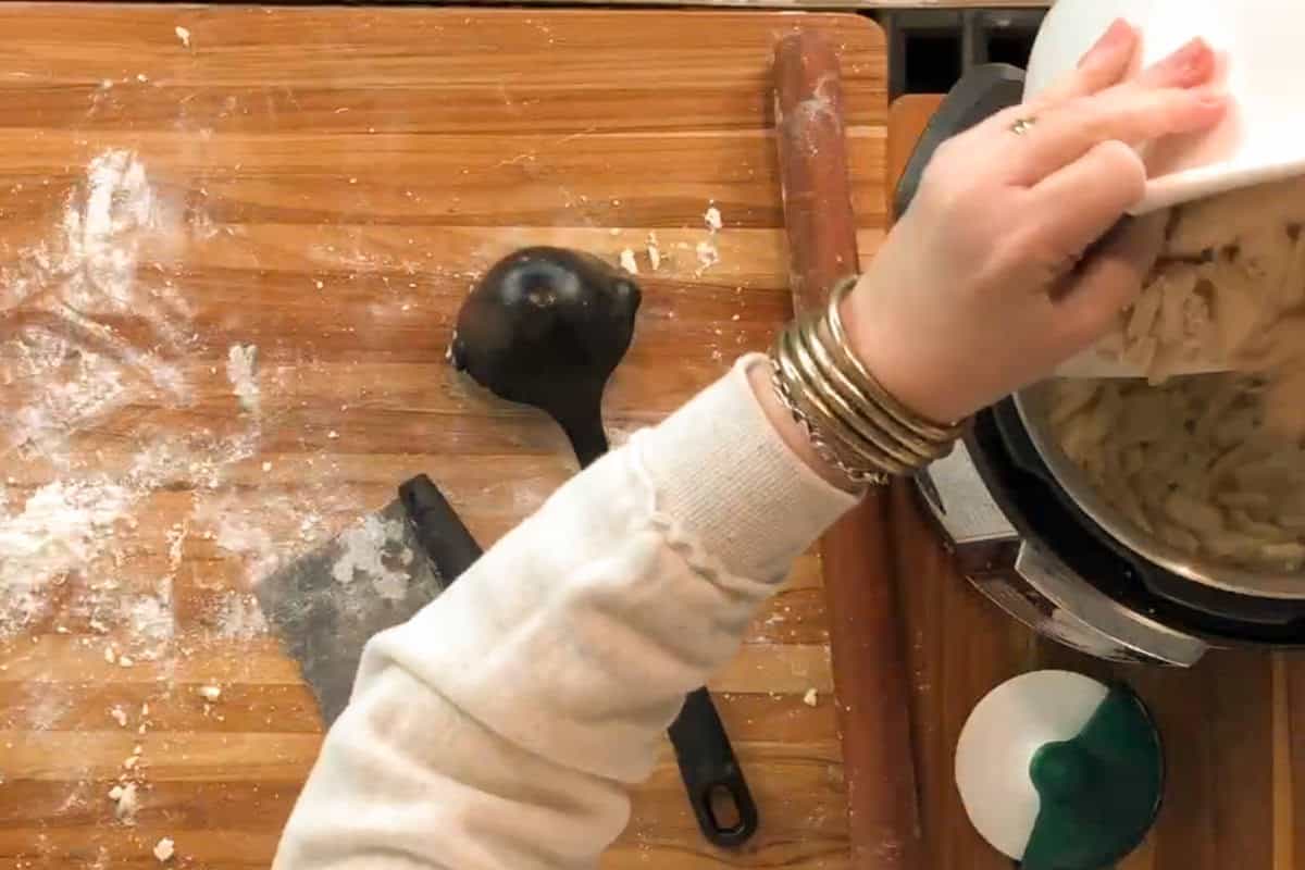 A person wearing a white sweater and several bracelets pours noodles from a white dish into a metal pot on a wooden surface, with flour, a ladle, and kitchen tools visible nearby.