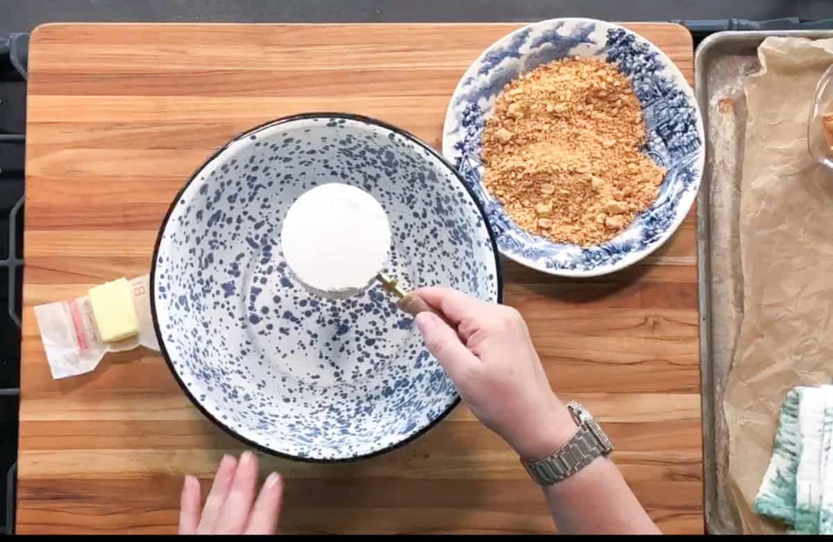 A person holds a measuring cup of sugar over a large white and blue speckled mixing bowl. Nearby are a bowl of graham cracker crumbs, a butter stick, and a baking tray lined with parchment paper.
