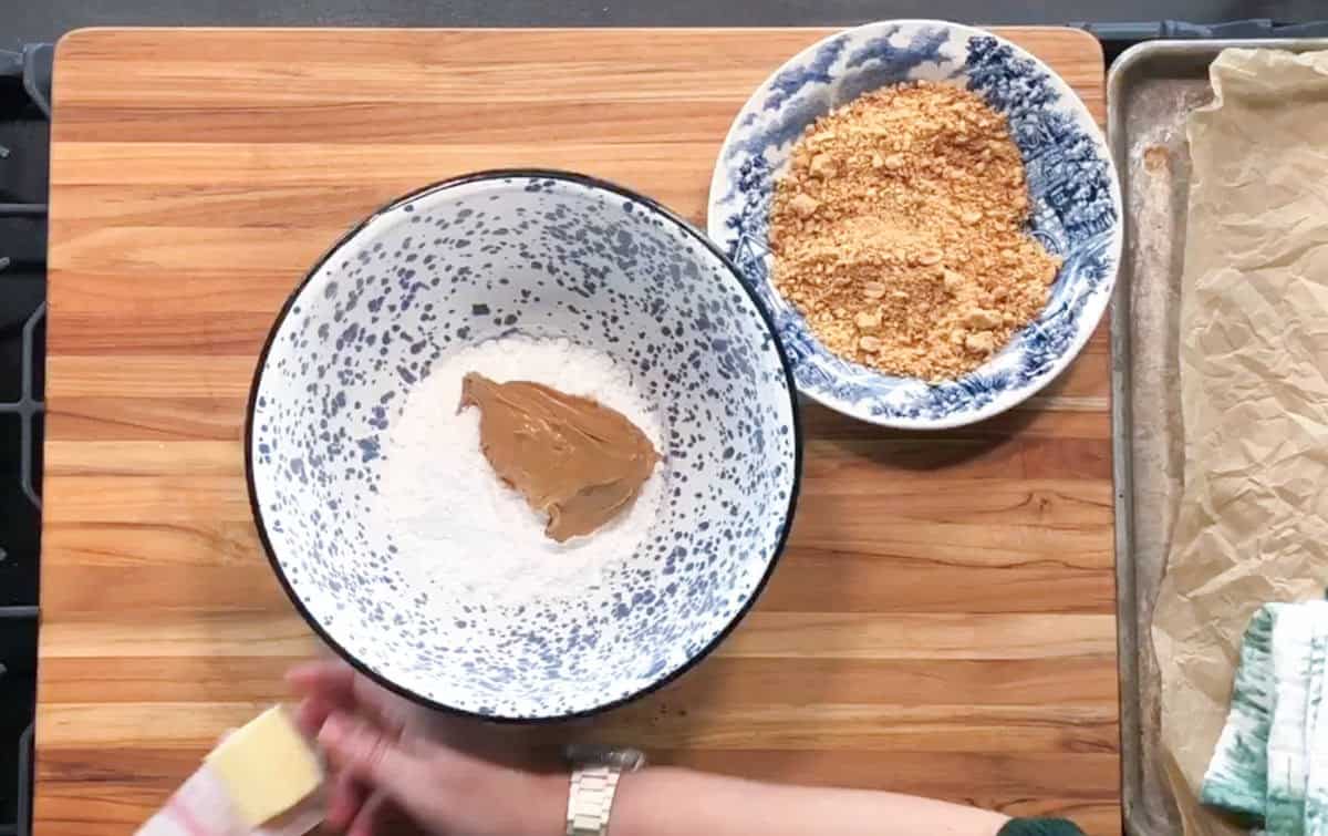 A hand holds a stick of butter over a mixing bowl containing flour and peanut butter. Next to it is a bowl of crushed crackers, all placed on a wooden cutting board with a baking sheet lined with parchment paper nearby.