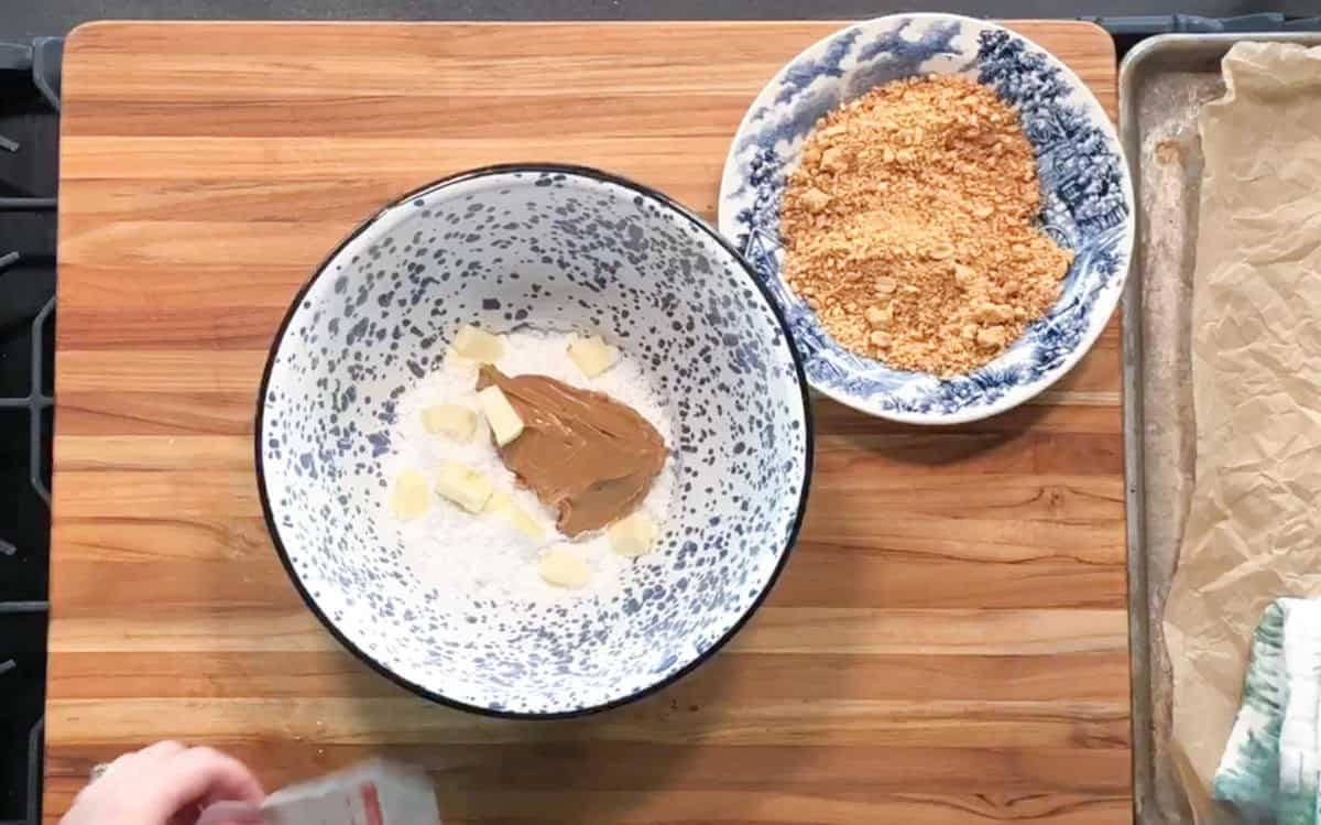 A mixing bowl with flour, small butter pieces, and peanut butter sits on a wooden surface next to a bowl of crushed graham crackers. A parchment-lined baking sheet and a hand holding a packet are partially visible.