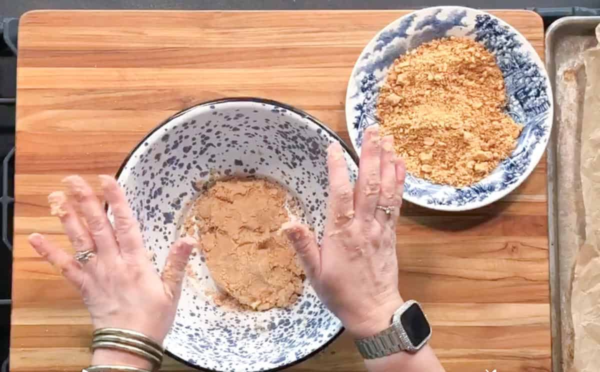 A person presses a crumbly mixture in a speckled bowl on a wooden surface. Nearby are a bowl of crushed crackers and a baking tray lined with parchment paper. The person wears a watch and several bracelets.