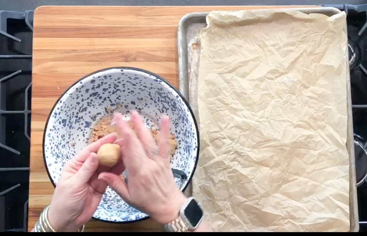 A person rolls dough into a ball over a speckled mixing bowl with dough inside. A baking tray lined with crumpled parchment paper sits to the right on a wooden cutting board.