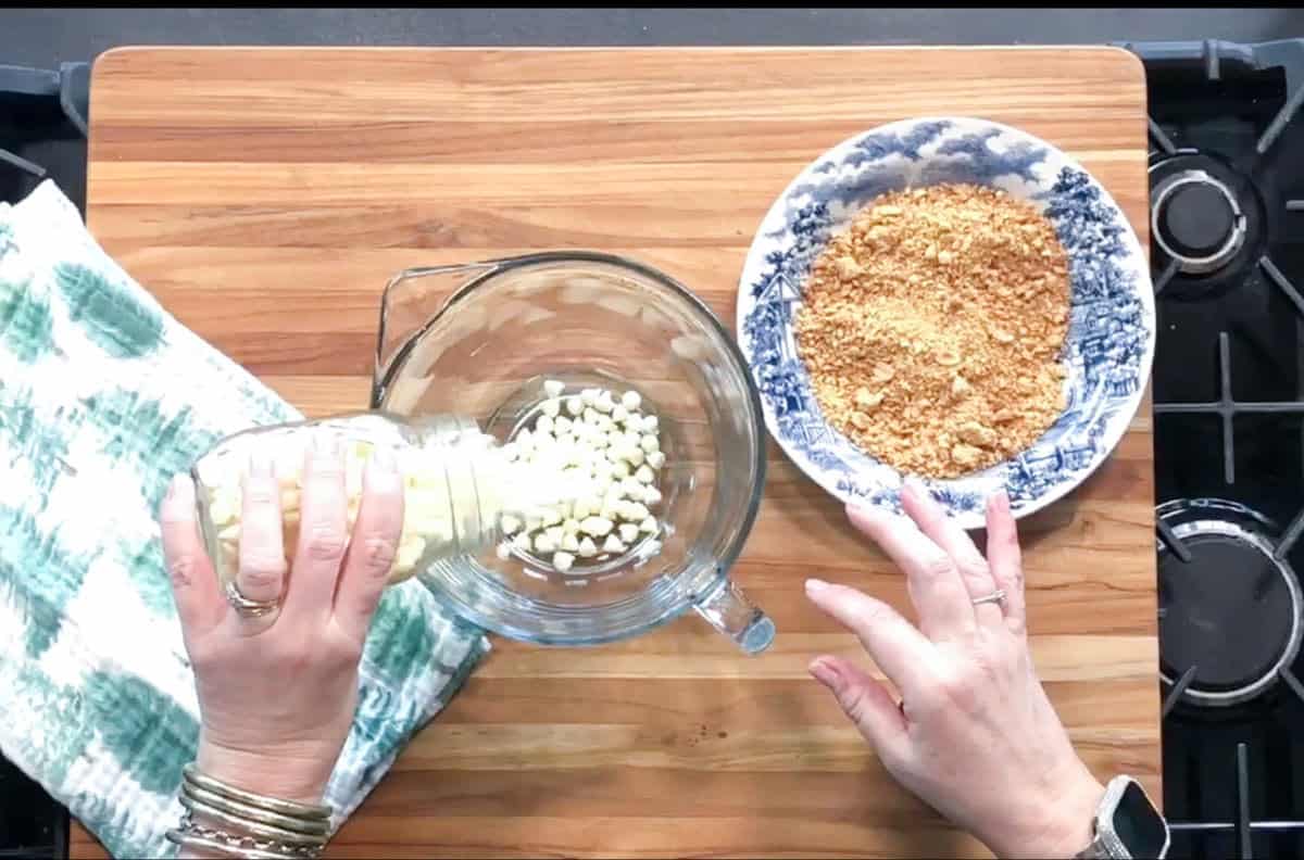 A person pours white chocolate chips from a jar into a glass mixing bowl on a wooden cutting board; a bowl of crushed cookies and a green and white towel are nearby.