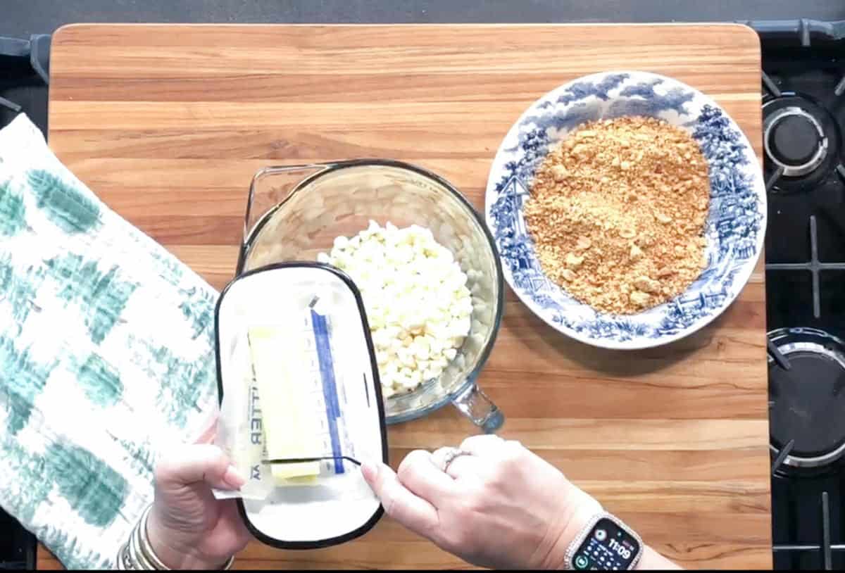 A person unwraps a stick of butter on a wooden cutting board next to a mixing bowl of white chocolate chips and a blue-patterned bowl filled with crushed graham crackers. A green and white towel is on the left.