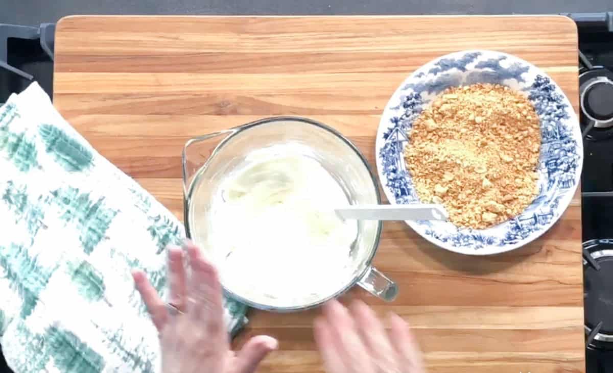 A person’s hands are visible near a glass bowl containing a white creamy mixture with a spoon, next to a blue and white bowl filled with crushed crackers, all placed on a wooden cutting board.