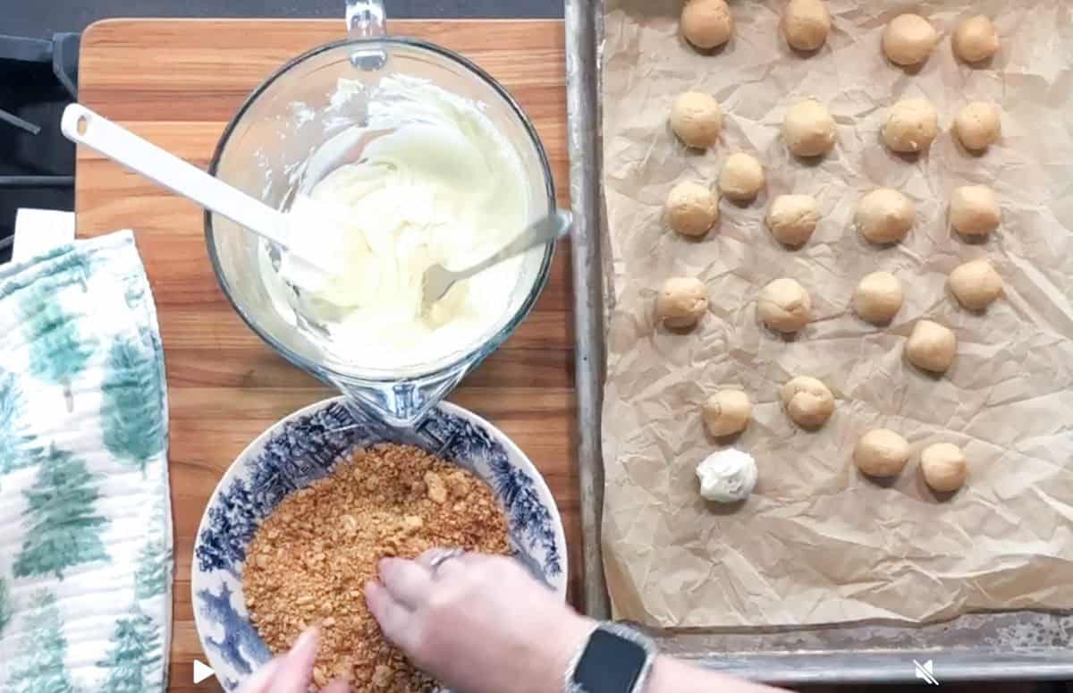 A person coats a small ball of dough in crushed crumbs over a blue and white bowl. A glass bowl of white mixture, baking sheet with dough balls, and oven mitt are on a wooden surface.
