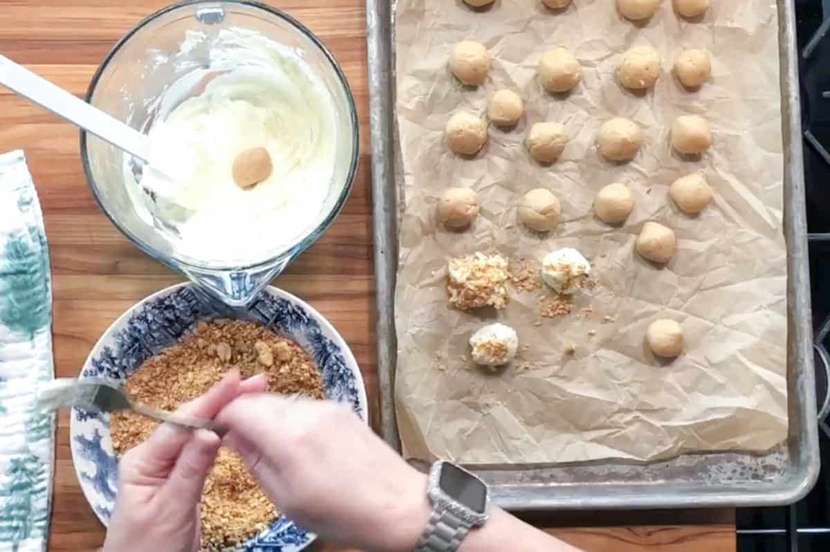 A person dips a round cookie into a bowl of white coating, rolls it in crushed nuts, and places it on a parchment-lined baking sheet with other coated and uncoated cookies. A mixing bowl and spoon are nearby.