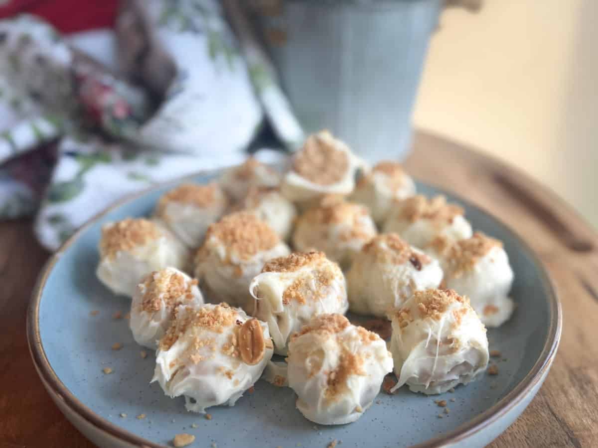 A plate of white chocolate-covered treats topped with crumbled pieces, arranged on a light blue ceramic plate on a wooden surface, with a patterned cloth and a metal container in the background.