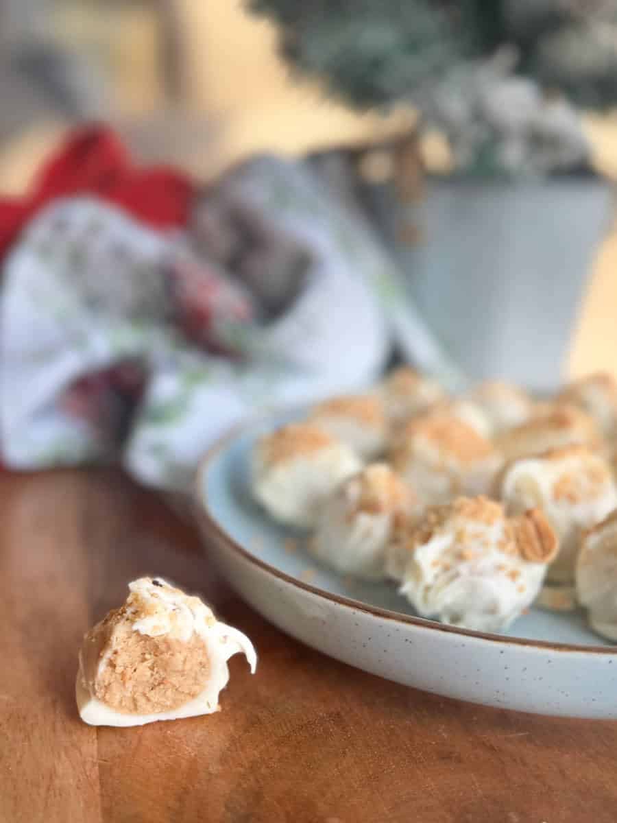 A close-up of a plate with several white chocolate-covered truffles, one truffle placed in front of the plate, on a wooden surface. A blurred cloth and potted plant are in the background.