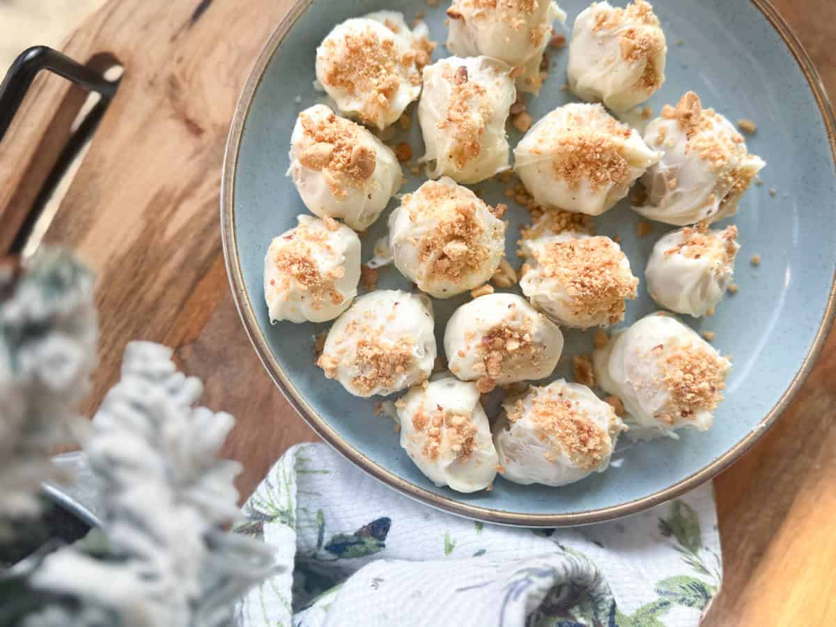 A blue plate holds several white chocolate-covered dessert balls topped with crushed cookie crumbs, placed on a wooden surface next to a floral-patterned cloth and a blurred plant.