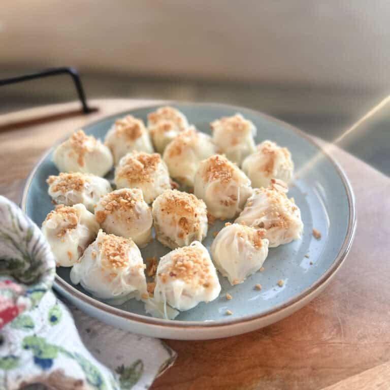 A blue plate holds several white chocolate-coated truffles topped with crushed nuts, arranged closely together on a wooden surface next to a patterned cloth.