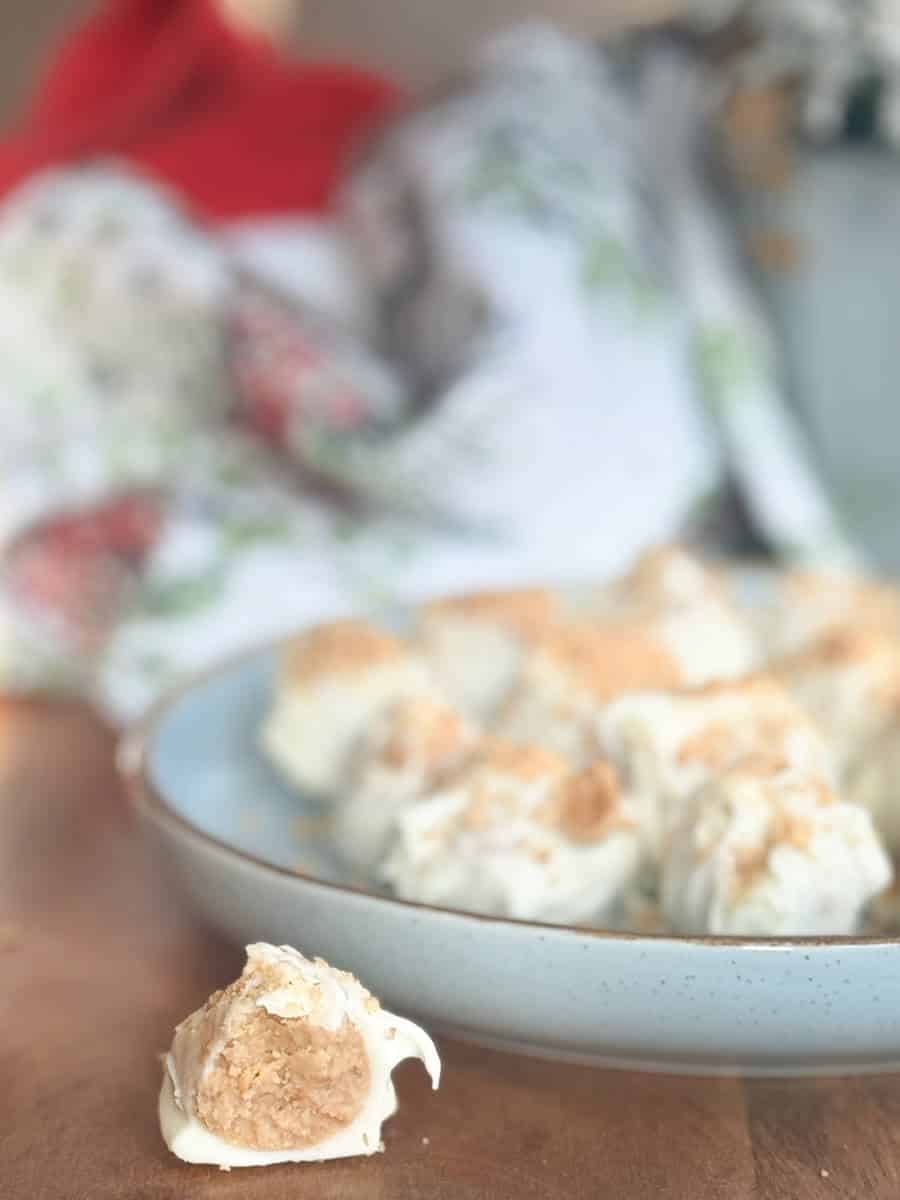 A close-up of a light blue plate with white chocolate-coated dessert bites, one bite in front of the plate showing a beige filling. The background is out of focus with a cloth and a red object visible.
