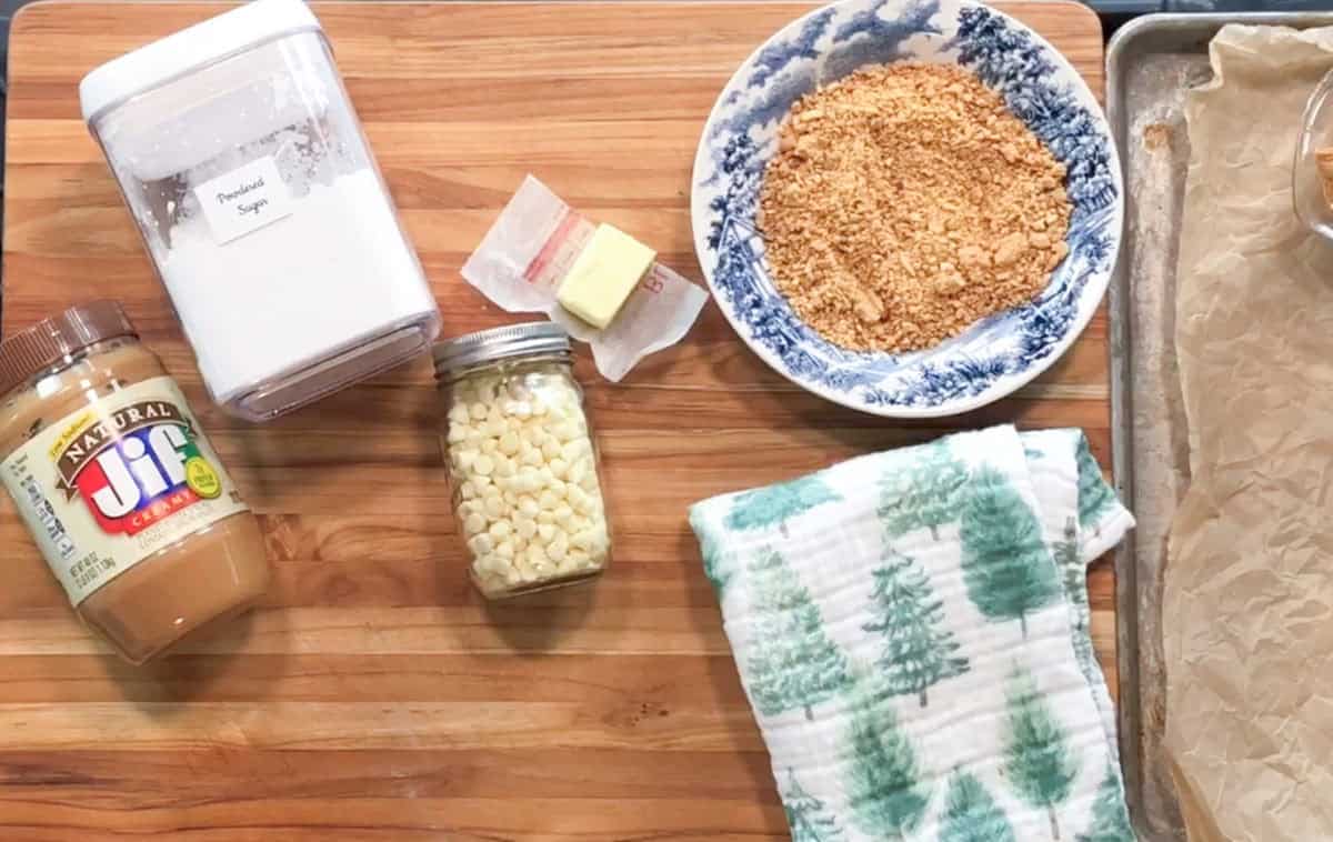 A jar of Jif peanut butter, a container labeled powdered sugar, a jar of white chocolate chips, a pat of butter, a bowl of crushed graham crackers, a green and white towel, and a parchment-lined baking sheet on a wooden surface.