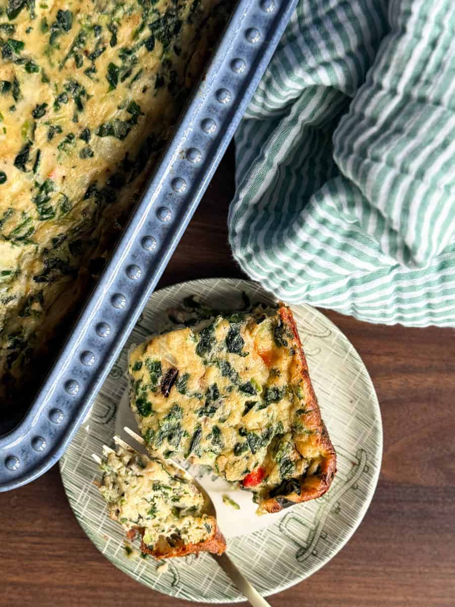 A slice of baked spinach and vegetable casserole sits on a plate with a fork. The casserole dish, mostly out of frame, is nearby. A green and white striped towel is next to the plate on a wooden surface.