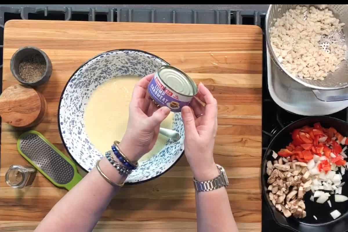 A person holds an opened can of chicken breast over a mixing bowl with beaten eggs. Nearby are a pepper container, a grater, and chopped vegetables, beans, and mushrooms on a stovetop.