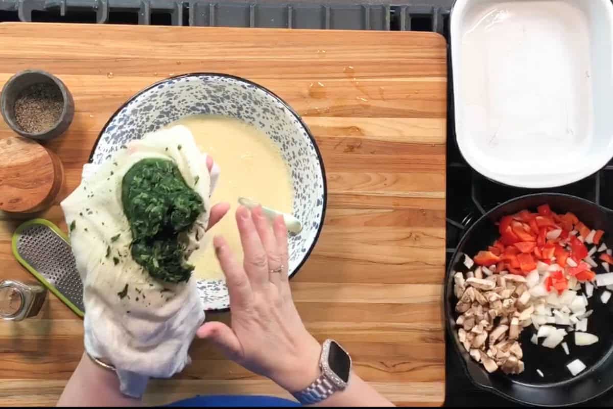 A person squeezes cooked spinach in a cloth over a bowl of beaten eggs. On the right, a pan holds chopped red bell pepper, onion, and mushrooms; an empty white baking dish sits nearby.