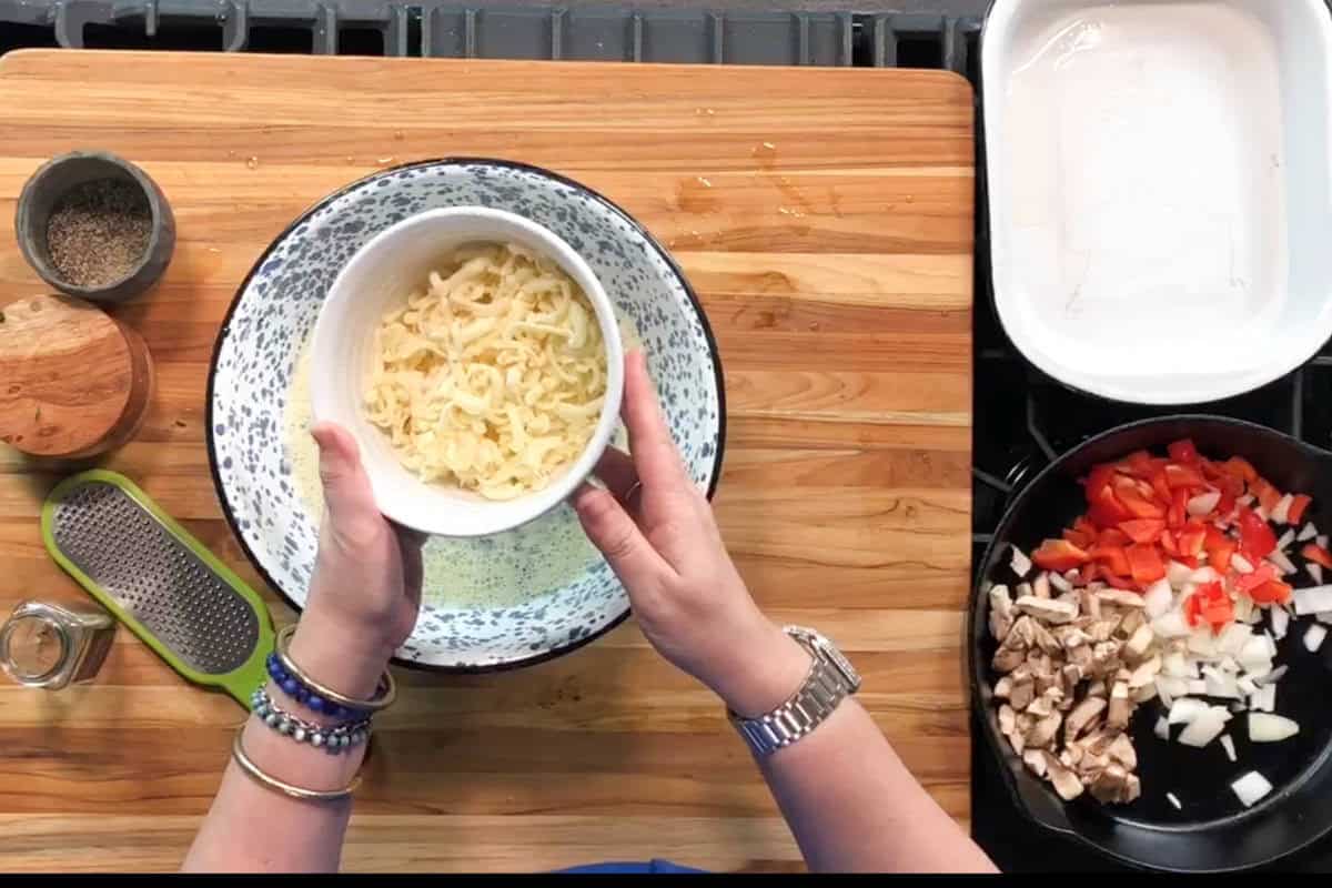 A person holds a bowl of shredded cheese over a mixing bowl on a wooden cutting board. Nearby are a small dish of pepper, a grater, a baking dish, and a pan with chopped onions, red peppers, and mushrooms.