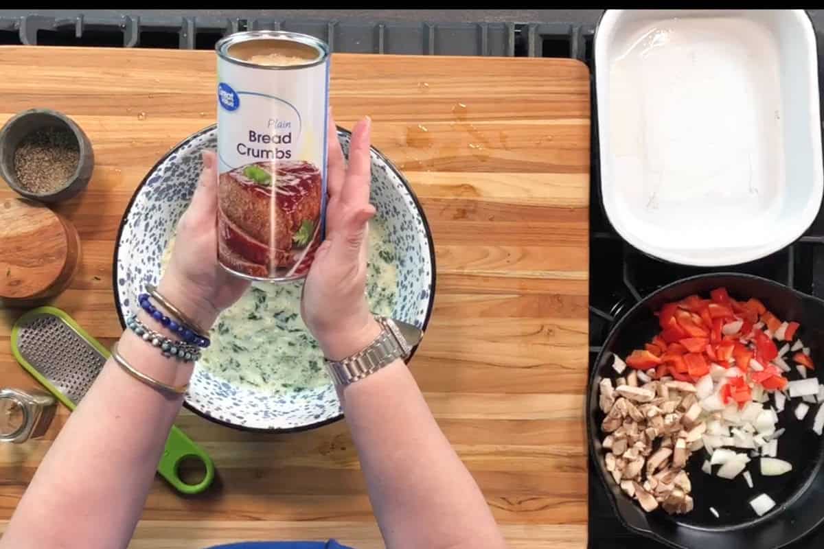 A person holds a can of plain bread crumbs above a mixing bowl on a wooden countertop. Nearby are measuring tools, seasonings, a baking dish, and a pan with chopped onions, red peppers, and mushrooms.