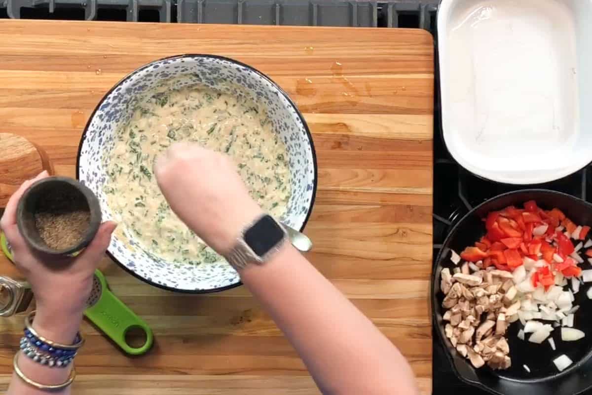 A person stirs a white speckled bowl of batter on a wooden cutting board. Their other hand holds a container of spices. Nearby, a pan contains chopped onions, red bell pepper, and mushrooms. A white dish sits beside it.