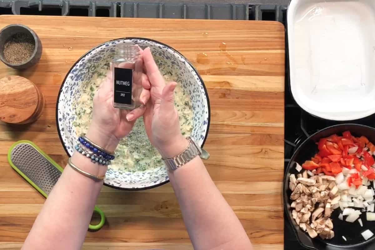 A person holds a jar labeled "nutmeg" over a bowl of mixture on a wooden counter. Nearby are a spice bowl, a grater, a baking dish, and a skillet with chopped mushrooms, onions, and red peppers.