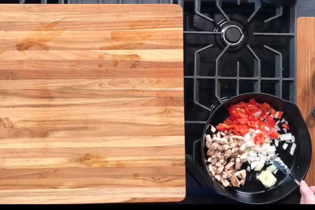 A black skillet on a gas stove contains chopped onions, red bell peppers, mushrooms, and a pat of butter. A hand is holding the skillet handle. Next to the stove is a large, empty wooden cutting board.