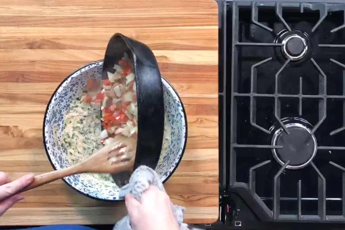 A person uses a wooden spoon to scrape sautéed vegetables from a black skillet into a bowl with a creamy mixture on a wooden countertop next to a black stovetop.