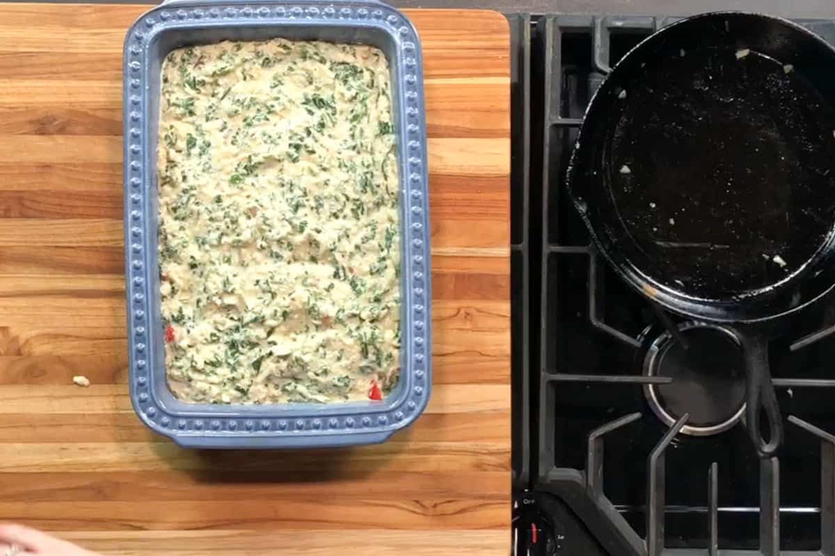 A blue rectangular baking dish filled with an uncooked mixture, likely containing chopped spinach, sits on a wooden countertop next to a black stove with a cast iron skillet.