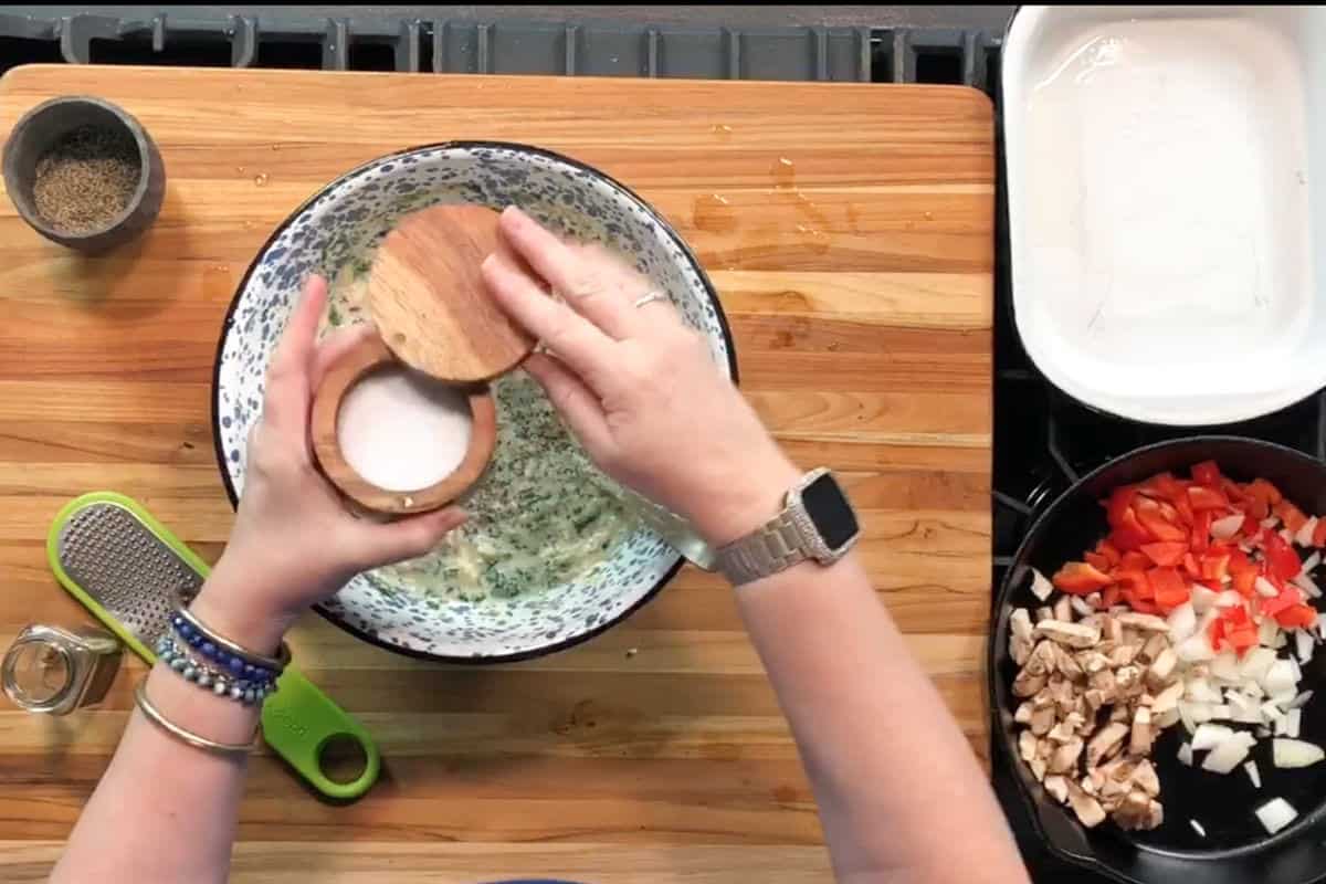 A person wearing a silver watch adds salt to a mixing bowl with herbs. On the counter are a grater, a small bowl of pepper, and a pan with chopped mushrooms, onions, and red peppers.