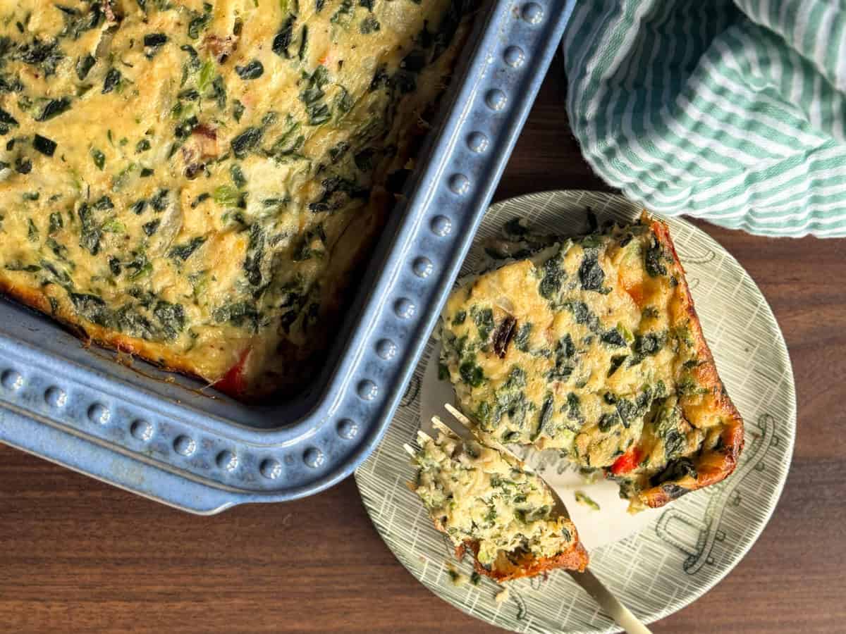 A baked casserole with visible leafy greens sits in a blue dish. Next to it, a square serving is placed on a plate with a fork, and a green and white striped cloth is nearby.