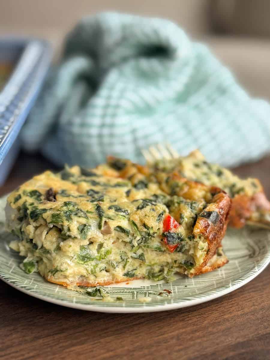 A slice of vegetable casserole with visible spinach, red peppers, and mushrooms sits on a green patterned plate. A fork rests in the casserole. A green and white checkered cloth is in the background.