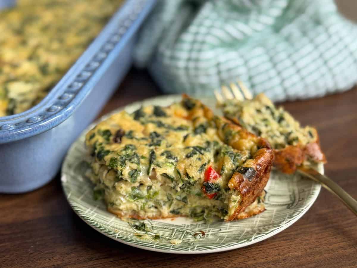 A slice of vegetable casserole with visible greens and red peppers sits on a patterned plate next to a fork, with a baking dish of casserole and a green checkered cloth in the background.