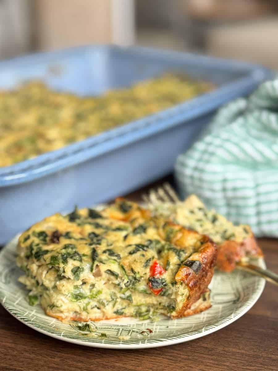 A slice of vegetable frittata sits on a green plate in the foreground, with a fork and a larger blue baking dish of frittata and a green striped cloth visible in the background.