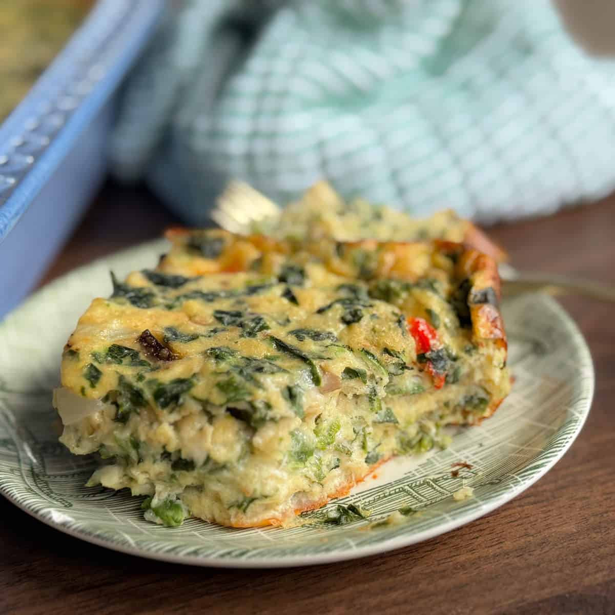 A close-up of a slice of vegetable casserole, featuring visible spinach, red bell peppers, and other vegetables, served on a decorative plate with a blue dish towel in the background.