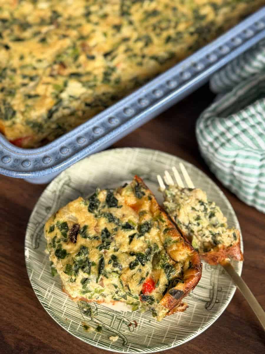 A slice of baked vegetable casserole with visible greens and red pieces sits on a plate with a fork. The rest of the casserole is in a rectangular baking dish nearby. A green checkered cloth is partially visible.