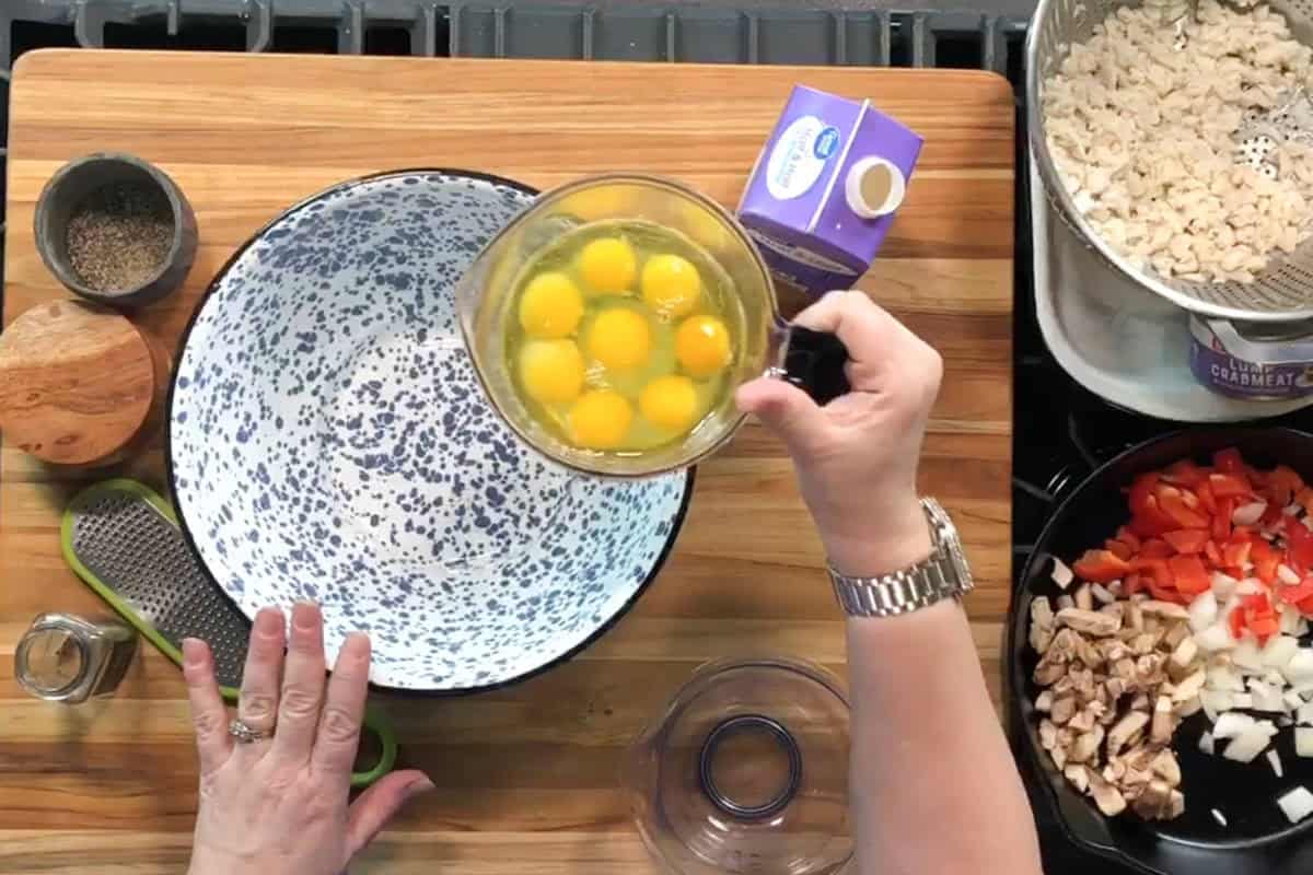 A person holds a glass measuring cup with cracked eggs over a large speckled mixing bowl on a wooden surface. Nearby are chopped vegetables, shredded cheese, a box of cream, and a small bowl of pepper.