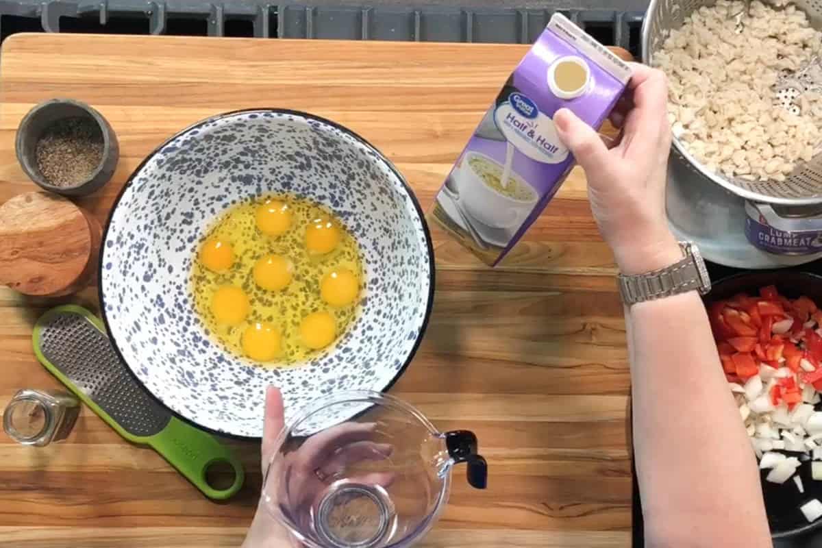 A person prepares a recipe in a kitchen. Six eggs are in a mixing bowl. The person holds a carton of half and half above the bowl, with various chopped ingredients and seasonings arranged on the wooden counter.