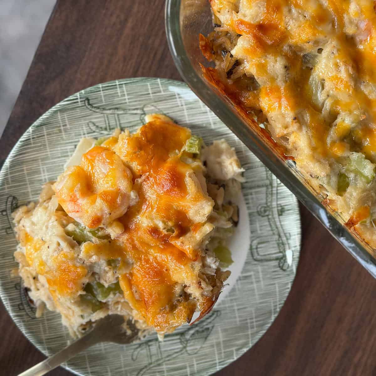A portion of cheesy casserole on a patterned plate with a fork beside a glass baking dish containing more of the baked casserole, set on a wooden surface.
