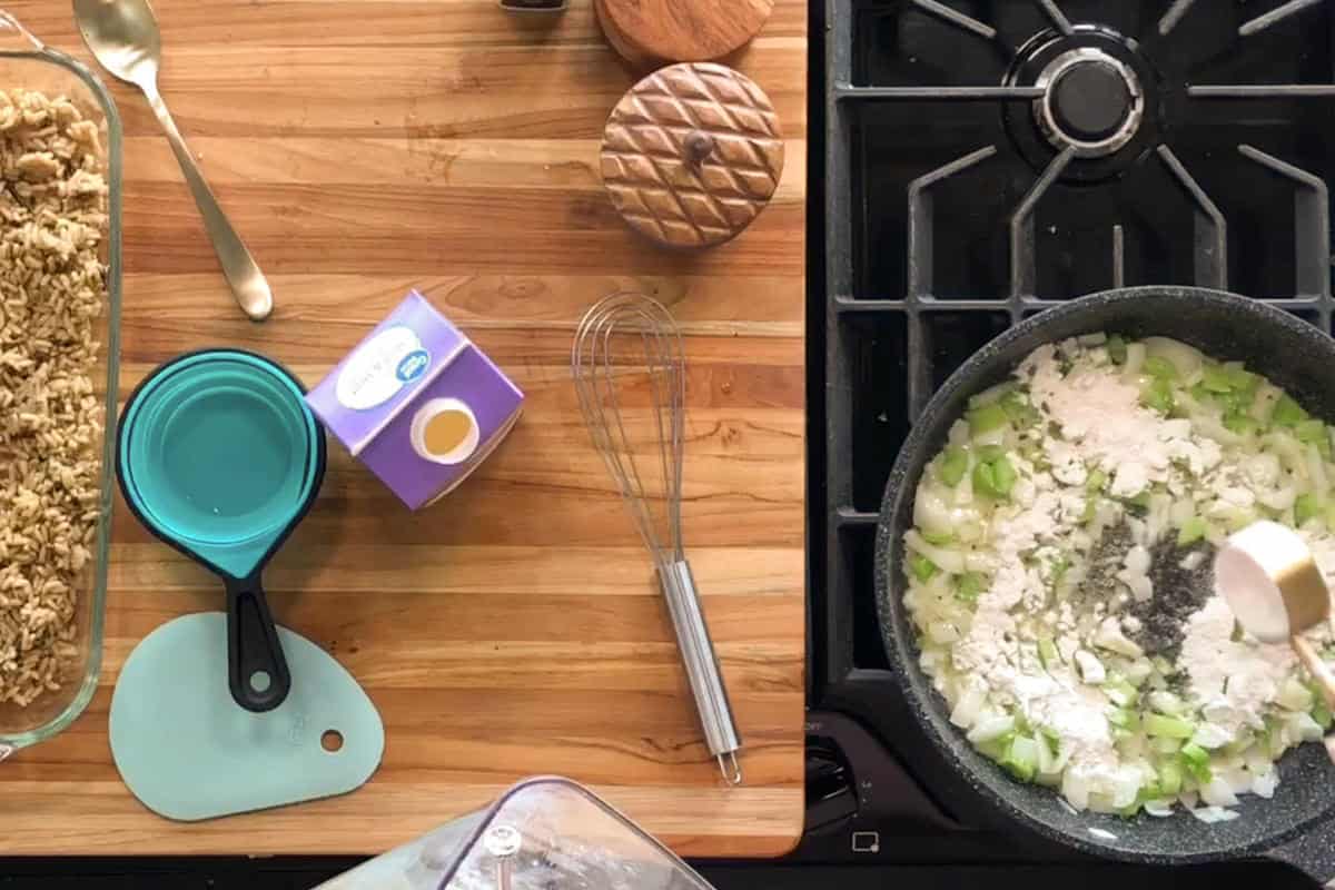 A glass baking dish with oat topping, measuring cups, a whisk, and a carton of cream sit on a wooden counter next to a stove where flour is being added to sautéed celery and onions in a black skillet.