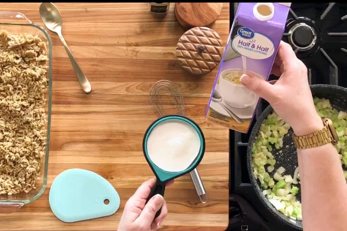 A person pours half & half from a carton into a green measuring cup above a stove with celery and onions cooking in a pan. A baking dish with oat crumble and kitchen utensils are on a wooden countertop.