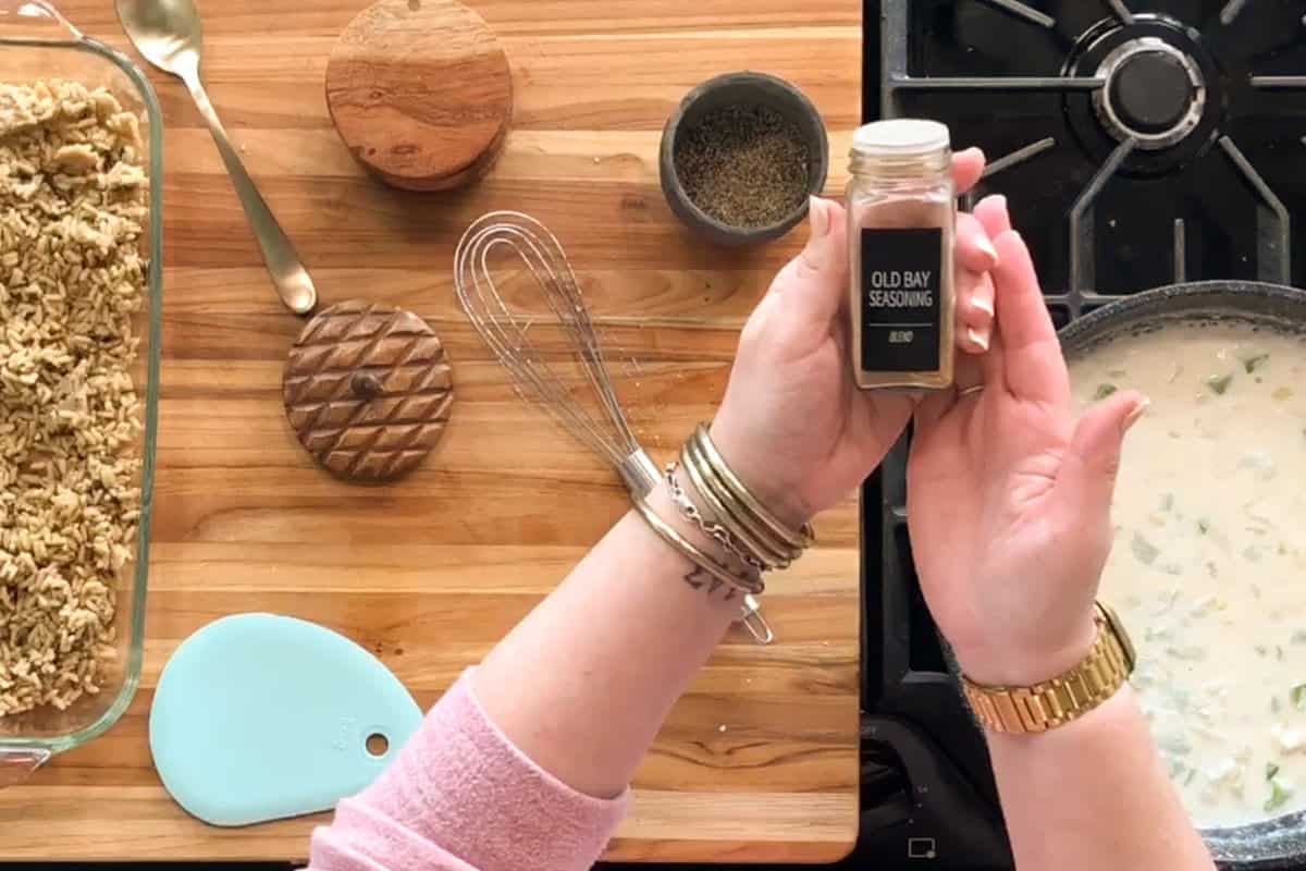 A person’s hands hold a jar of Old Bay seasoning over a wooden kitchen counter with a whisk, spice containers, a blue spatula, an oat topping dish, and a stove with a pot of creamy mixture.