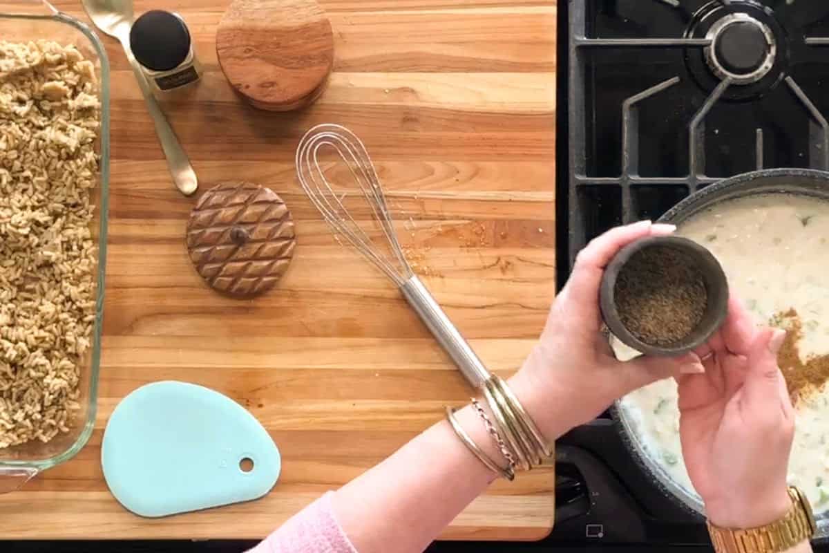 A person sprinkles seasoning into a saucepan of creamy mixture on a stove. On the wooden counter are a whisk, a blue scraper, two round containers, a small spice jar, and a dish with an oat topping.