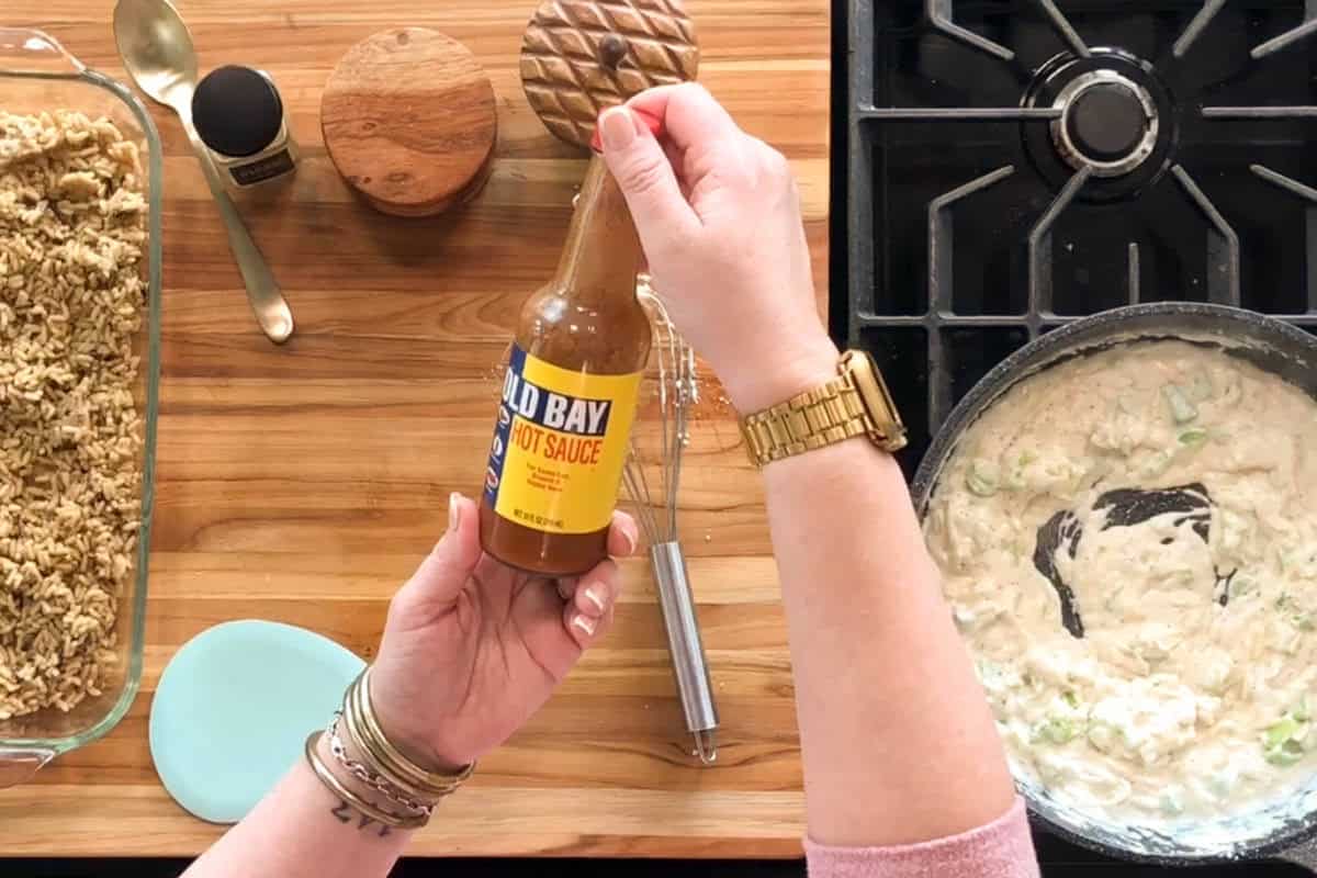 A person holding a bottle of Old Bay Hot Sauce above a wooden countertop with a baking dish of crumble, kitchen utensils, and a stove with a creamy mixture in a pan.