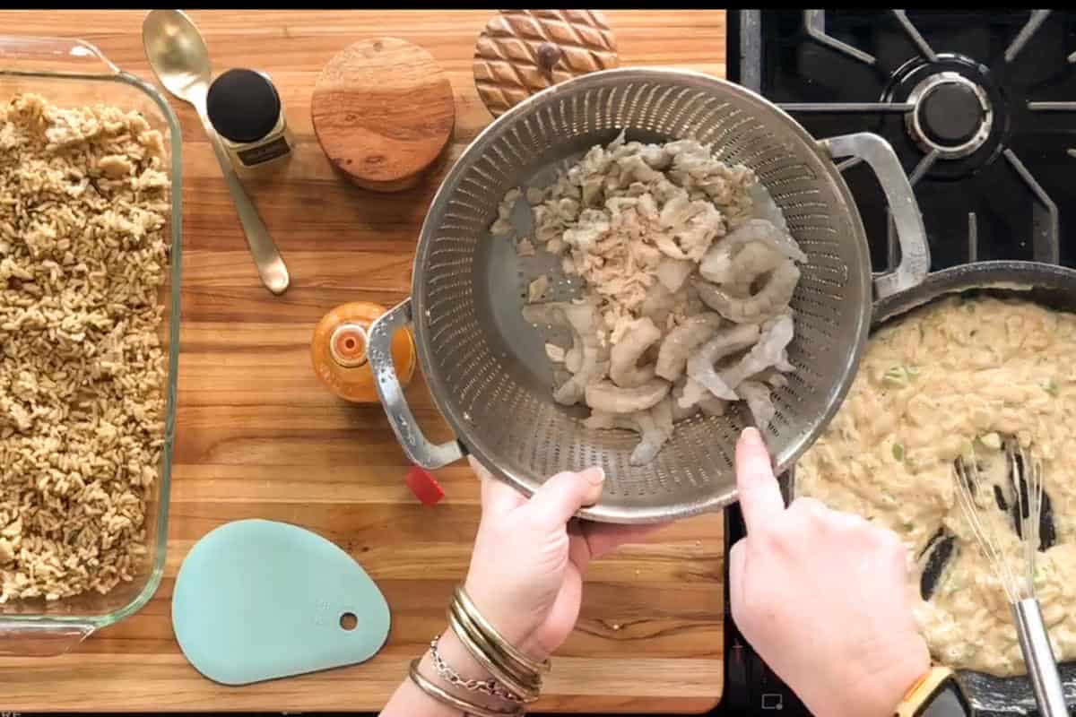 A person holds a colander of raw shrimp over a skillet of creamy sauce on a stove. On the left, a baking dish with a crumb topping and various kitchen tools are on a wooden countertop.
