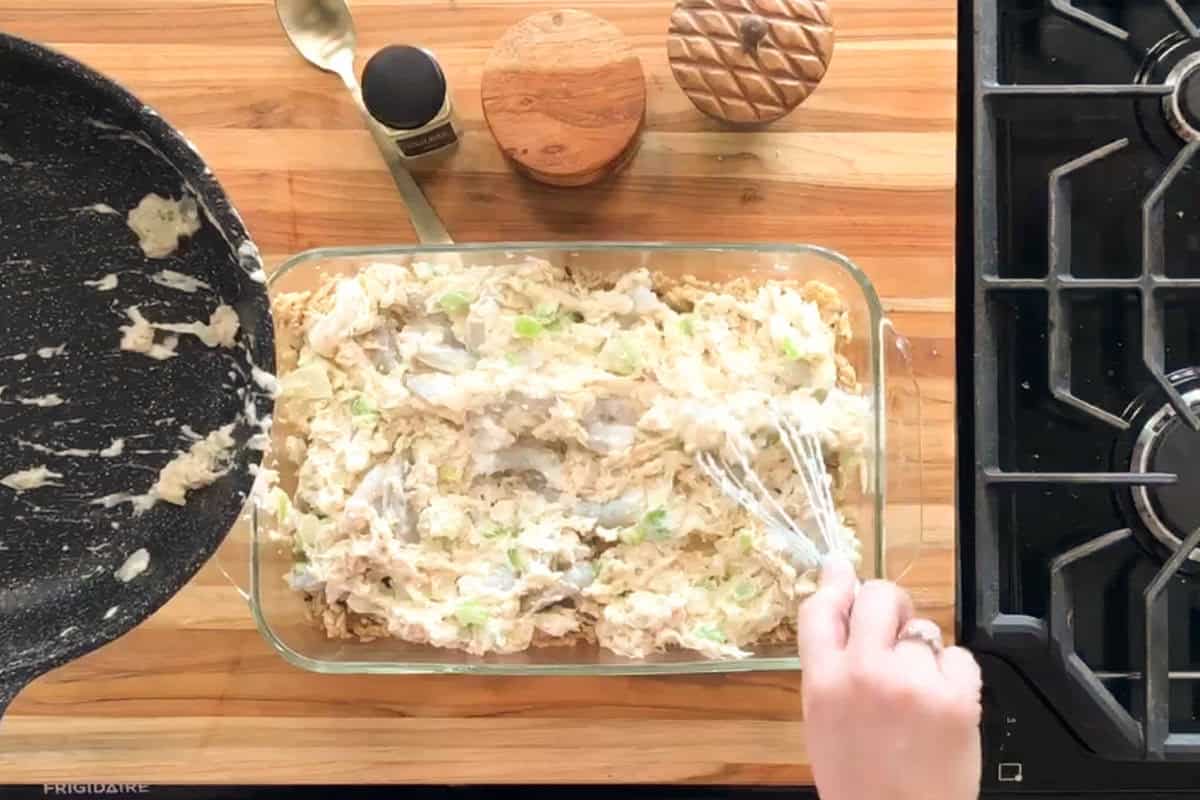 A person spreads a creamy casserole mixture into a glass baking dish on a wooden countertop beside a stove, with a black skillet, salt and pepper shakers, and a hand holding a whisk.