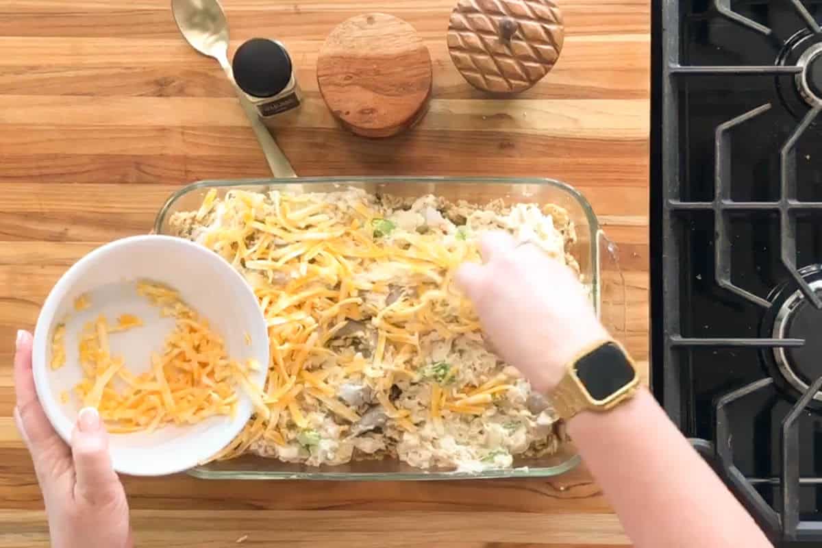 A person sprinkles shredded cheese from a white bowl onto a casserole in a glass baking dish on a wooden counter near a stove. Seasoning containers and a gold spoon are also on the counter.