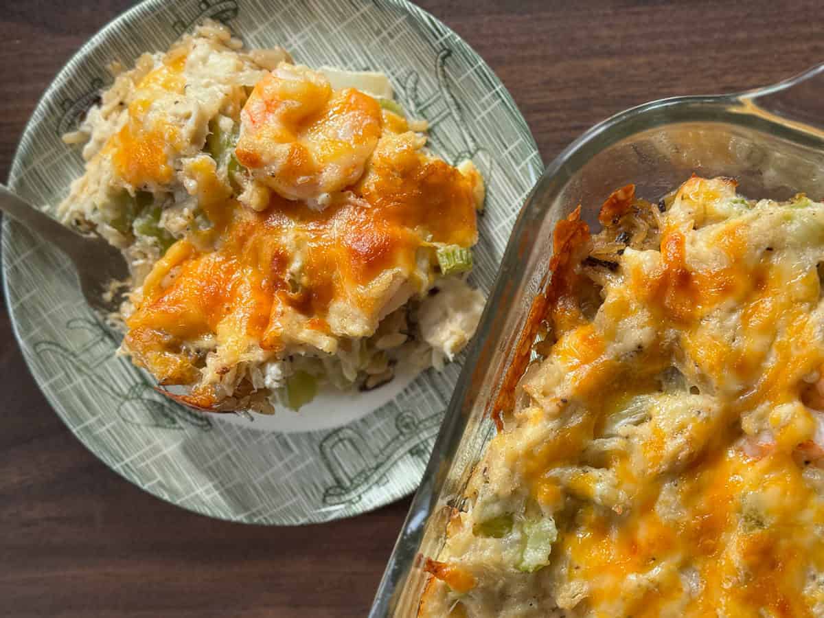A plate and a glass baking dish with baked casserole topped with melted cheese, showing chunks of chicken, rice, and vegetables on a brown tabletop.