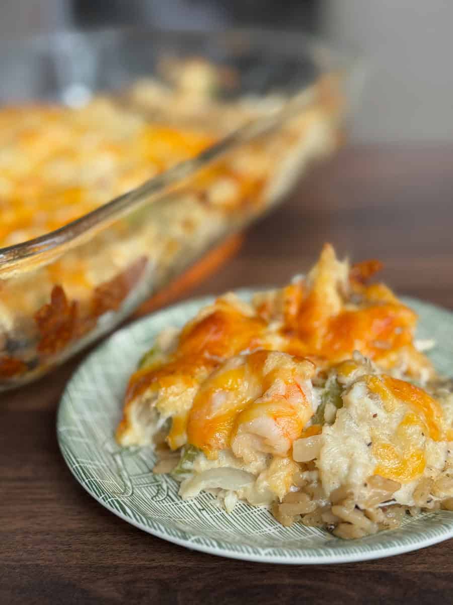 A close-up of a plate with a serving of cheesy casserole containing rice and shrimp, sitting in front of a glass baking dish with more of the same casserole. The casserole is topped with melted cheese.